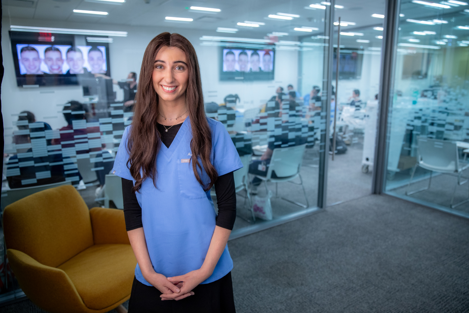 Ava Zelefsky smiling and wearing dental scrubs in front of large windows of dental classroom.