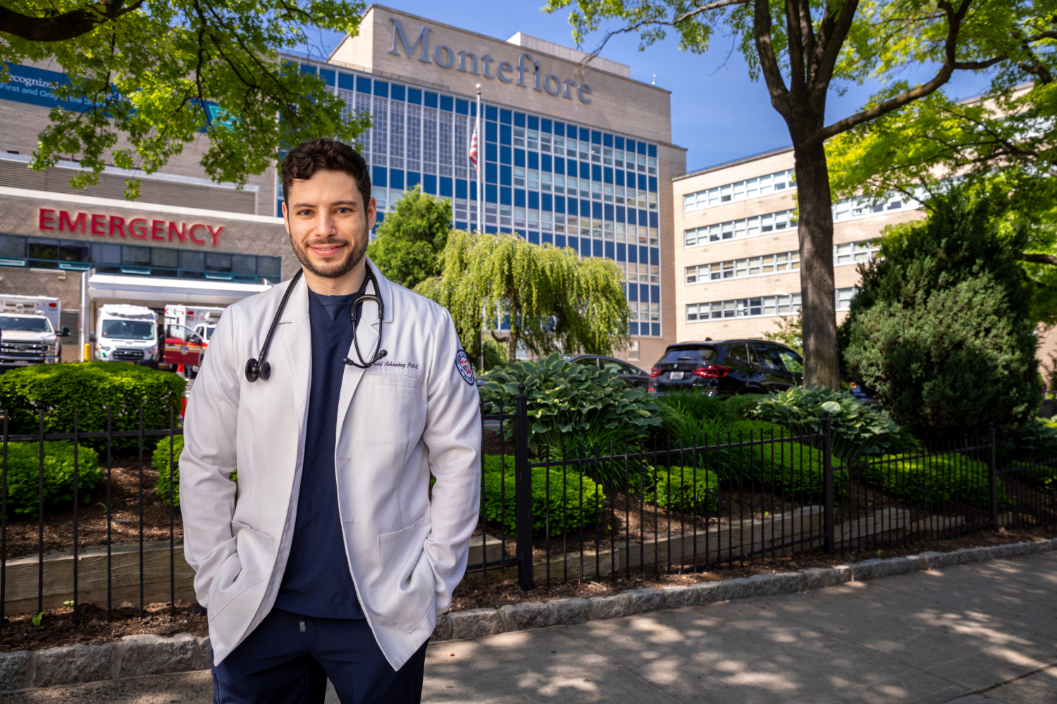 Dovid Schoenberg, wearing a white coat and a stethoscope wrapped around neck, posing outside of Montefiore Medical Center.