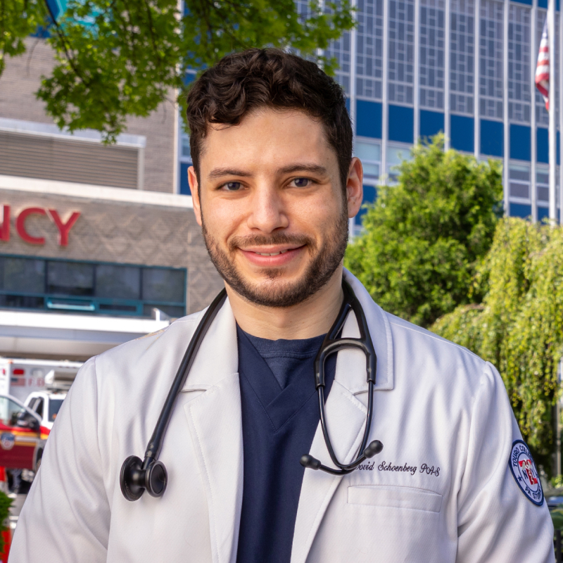 Dovid Schoenberg, wearing a white coat and a stethoscope wrapped around neck, posing outside of Montefiore Medical Center.