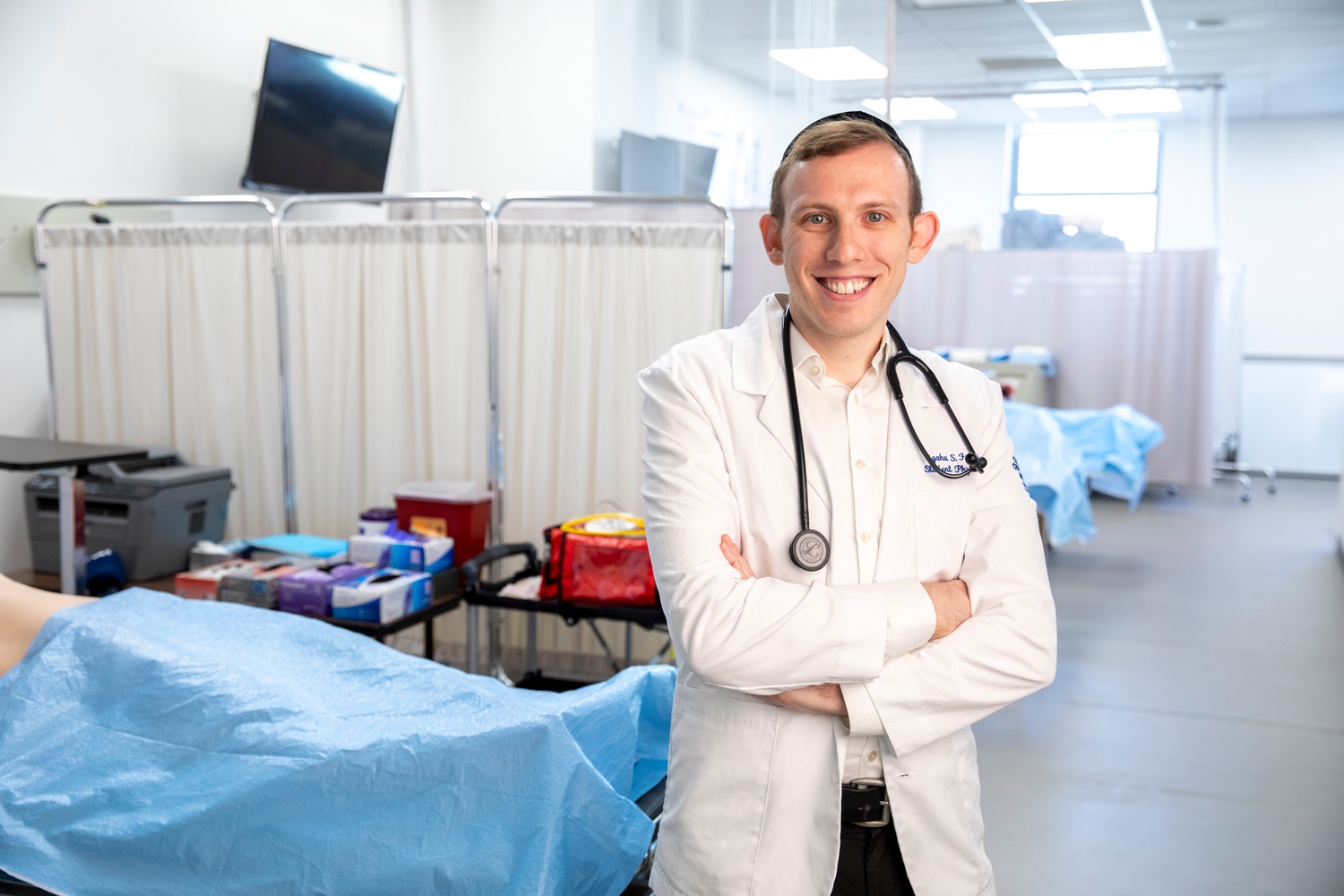 Eliyahu Freund smiling while wearing white coat and stethoscope around neck in TouroCOM practice clinic