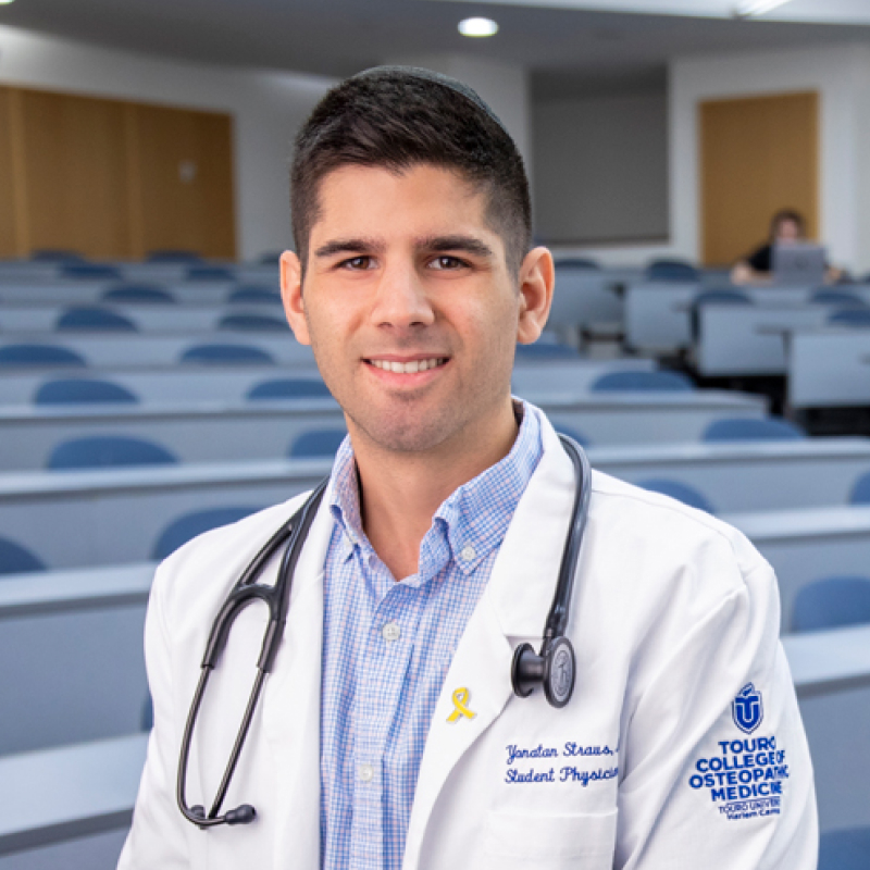 Yonatan Straus smiling while wearing a white coat and a stethoscope in lecture hall