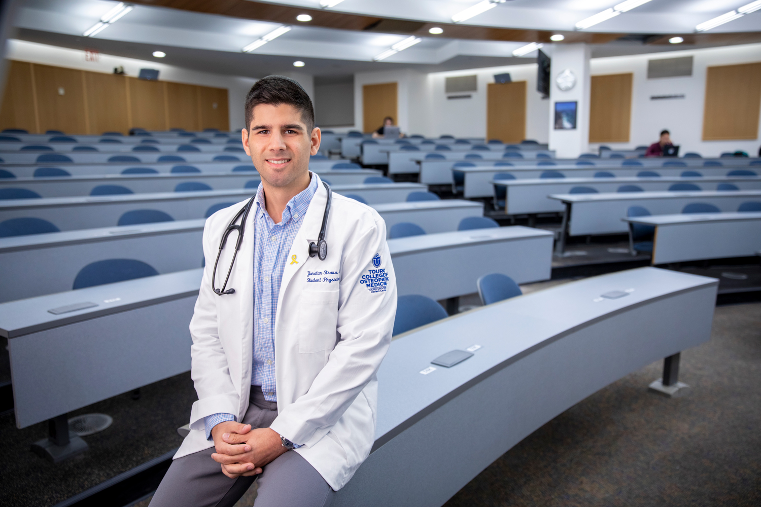 Yonatan Straus smiling while wearing a white coat and a stethoscope in lecture hall